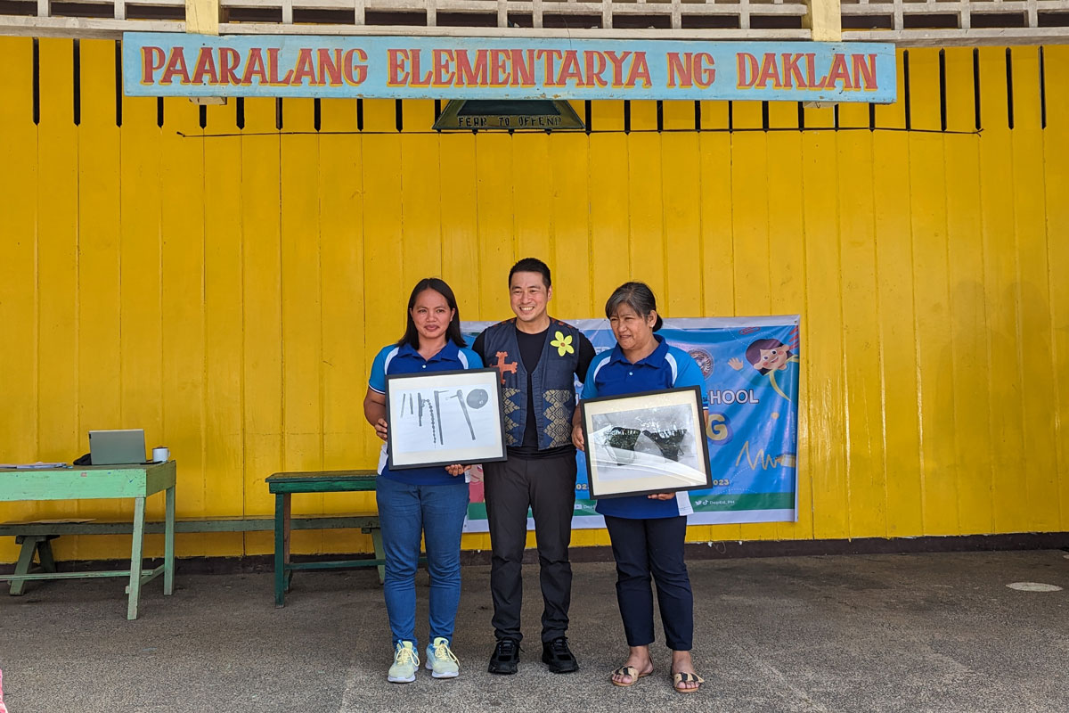 Three people holding framed photographs in front of a school in the Philippines.