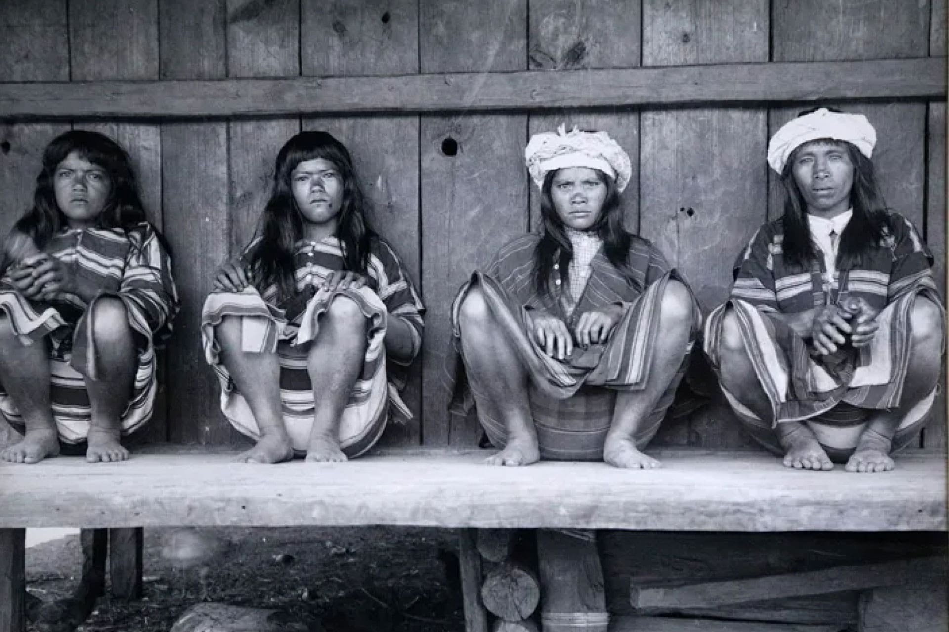 Four indigenous persons seated against a wooden wall.