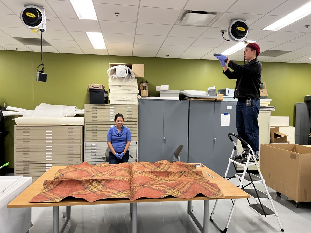 A person standing on a stool photographing a woven mat and another person seated.